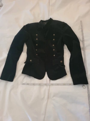 Svart sammetskavaj med guldknappar - Stilren svart sammetskavaj med dubbelknäppning och dekorativa guldknappar framtill och på ärmarna. Figurnära modell med markerad midja och två fickor fram. Perfekt för dig som vill ha en elegant och trendig look.