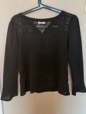 Svart blus med spets från Only - Stilren svart blus från Only med v-ringning och spetsdetaljer över axlar och bröst. Blusen har långa ärmar och är tillverkad i ett lätt, flowy material som ger en snygg siluett. Perfekt att matcha med jeans eller kjol för en trendig look.
