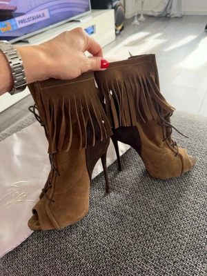 Bruna mocka stövletter med fransar - Säljer ett par bruna stövletter från Giuseppe Zanotti i mjuk mocka. Skorna har höga smala klackar, öppen tå och snörning framtill. Dekorativa fransar runt skaftet ger en cool boho-vibe. Perfekta för dig som vill sticka ut med din stil.