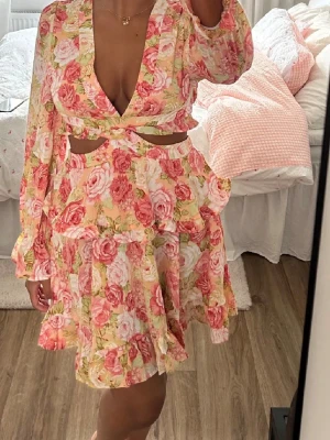 Blommig kortklänning med volanger - Blommig kortklänning i rosa, vit och grön med volangdetaljer, djup v-ringning och cut-outs i midjan. Klänningen har långa ärmar med resår vid handlederna och knyt i ryggen. Perfekt för sommarens alla tillfällen.