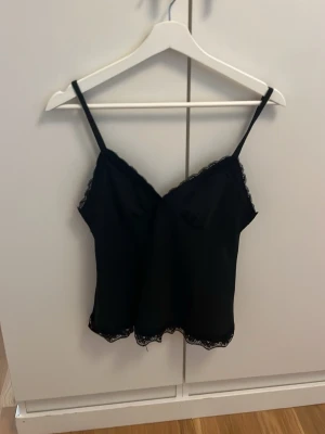 Svart linnetopp med spetsdetaljer - Snygg svart linnetopp med tunna axelband och spetsdetaljer längs urringning och nederkant. Perfekt att styla lager på lager eller bära som den är för en trendig look.