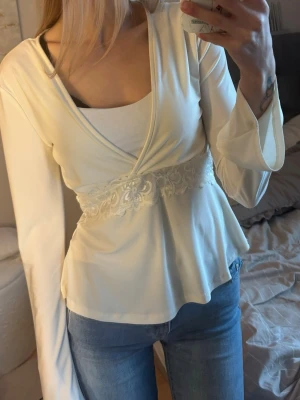 Vit omlottblus med spetsdetalj - Säljer en vit omlottblus med långa, utsvängda ärmar och vacker spetsdetalj i midjan. Blusen har en feminin v-ringning och är tillverkad i ett mjukt, lätt material som ger ett snyggt fall. Perfekt att styla med jeans eller kjol.