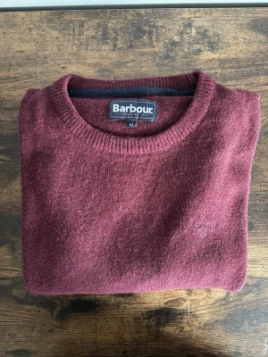 Barbour Tröja - Barbour stickad tröja i storlek M. Klassisk modell i vinröd färg med diskret logga på bröstet. Clean och tidlös design som passar perfekt till casual/old money fits. Använd men fortfarande i bra skick, mindre tecken på användning finns.