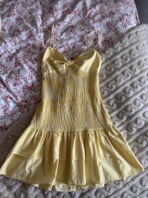 Gul rutig kortklänning med rosett - Söt gul och vit rutig kortklänning med smala axelband och volangkant. Klänningen har en stor rosett framtill och omlott-detalj vid bysten. Perfekt för varma dagar och ger en fräsch sommarkänsla. Lite i storlek därför säljer jag som en Medium 