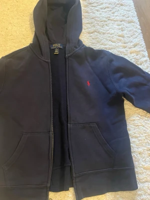 Marinblå hoodie från Polo Ralph Lauren - Klassisk marinblå hoodie från Polo Ralph Lauren med dragkedja, huva och kängurufickor. Ikoniska röda loggan broderad på bröstet. Tillverkad i mjuk bomullsblandning som är perfekt för chill dagar eller när du vill ha en clean look.