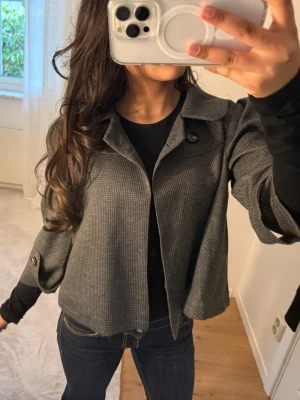 Grå rutig overshirt - Snygg grå overshirt med diskret rutigt mönster och stora knappar framtill samt på ärmarna. Jackan har en klassisk krage och en loose passform som ger en avslappnad vibe. Perfekt att slänga över en hoodie eller t-shirt för en cool lager-på-lager-look.