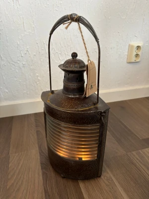Antik metalllykta med glasruta - Säljer en gammaldags lykta i mörk metall med rundad glasruta framtill och dekorativt handtag. Lyktan har ett detaljerat lock med ventilationshål och en etikett i snöre. Perfekt som inredningsdetalj för dig som gillar vintage och rustik stil.