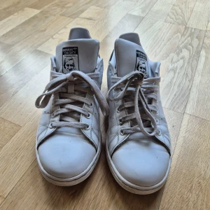 Adidas Stan Smith vita sneakers - Klassiska vita Adidas Stan Smith sneakers med lågt skaft och svarta detaljer på hälen. Skorna har perforerade ränder på sidorna, vita snören och är tillverkade i slätt läder. Ikonisk logga på plösen och hälen. Perfekta för en clean och stilren look.