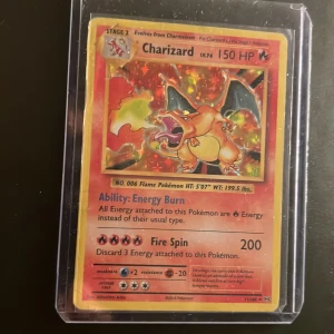 Charizard Pokémonkort Base Set - Säljer ett Charizard holografiskt Pokémonkort från Base Set. Kortet har en färgstark design med Charizard i rött och orange, och är innesluten i ett genomskinligt plastfodral. Perfekt för samlare eller Pokémonfans.