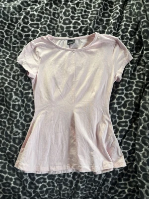 Ljusrosa peplum topp från Gina Tricot - Supersöt ljusrosa topp från Gina Tricot med rund halsringning och korta ärmar. Modellen har en lätt utsvängd passform nedtill och är gjord i mjuk bomull. Aldrig använd. Tvättad 1 gång. 