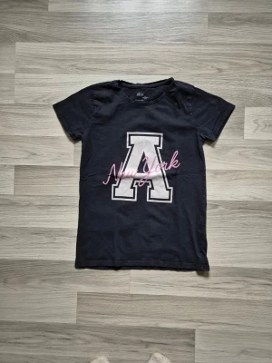 Svart t-shirt med New York-tryck - Snygg svart t-shirt från Ellos med stort vitt A och rosa texten 'New York' på bröstet. Klassisk rund halsringning och korta ärmar. Perfekt till jeans eller shorts för en avslappnad look.