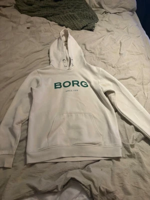 Vit hoodie från BORG - Säljer en vit hoodie från BORG med grön logga och texten 'SINCE 1984' på bröstet. Hoodien har en klassisk känguruficka framtill och ribbade muddar vid ärmslut och nederkant. Perfekt för en avslappnad och sportig stil.