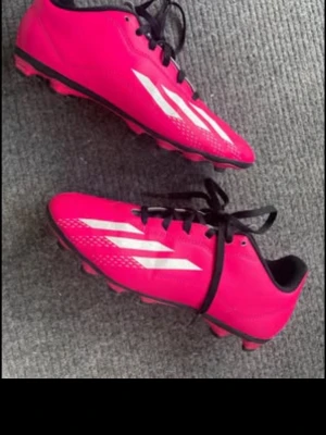 Rosa Adidas fotbollsskor med dobbar - Säljer ett par snygga rosa Adidas fotbollsskor med klassiska vita ränder på sidorna. Skorna har svarta snören, låg profil och fasta dobbar för spel på gräs. Ovandelen är i syntetmaterial som ger bra bollkontroll och passform. Använd fåtal ggr