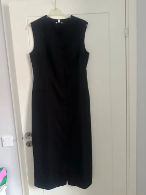 Långklänning stl L slim fit  - Klassisk svart långklänning i minimalistisk design. Klänningen är ärmlös med rund halsringning och rak passform. Perfekt för dig som gillar stilrena plagg. Materialet känns slätt och har en lätt struktur. Har knappar hela vägen. 