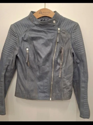 Grå biker skinnjacka från primeboots - Snygg grå skinnjacka i bikerstil med asymmetrisk dragkedja, ståkrage med tryckknapp och vadderade detaljer på axlar och ärmar. Två snedställda fickor med dragkedja framtill och coola sömmar som ger jackan en edgy look.