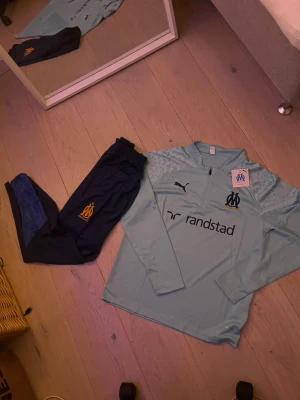 Marseille träningsset från Puma - Snyggt träningsset från Olympique de Marseille och Puma. Tröjan är ljusblå med halv dragkedja, klubbmärke och sponsortryck. Byxorna är mörkblå med klubbmärke och elastisk midja. Perfekt för fotbollsträning eller chill. Materialet är funktionspolyester.