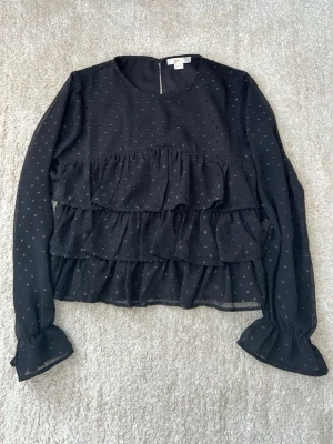 Svart volangblus med prickar - Supersöt svart blus från H&M med små prickar och flera volanglager framtill. Blusen har långärmade ärmar med volang vid ärmslut och är gjord i ett tunt, transparent tyg som ger en luftig känsla. Perfekt för dig som gillar trendiga detaljer och en feminin look.