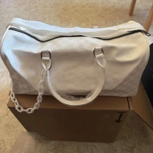 Vit lyxig weekendbag med rutmönster - Stilren vit handväska i rektangulär form med diskret rutmönster. Väskan har två handtag, silverfärgade metalldetaljer och en vit kedjedetalj i plast på sidan. Dragkedja upptill och tillverkad i ett slätt material som ger en modern look.      Mått: 43cm lång 21cm hög 18cm djup