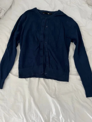 Mörkblå kofta från 157 - Mörkblå kofta från 157 med rund halsringning och knäppning framtill. Långärmad modell i mjukt material, perfekt att slänga på sig över en t-shirt eller linne. Klassisk och enkel design som funkar till det mesta.