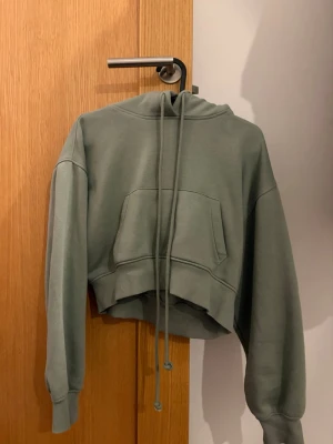 Grön cropped hoodie med huva - Snygg grön cropped hoodie med huva och långa dragsnören. Hoodien har en stor magficka framtill och ribbade muddar vid ärmslut och nederkant. Perfekt för en avslappnad och trendig look.
