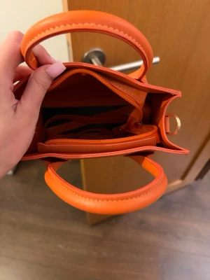 Mini orange handväska med handtag - Snygg liten handväska i stark orange färg med två rundade handtag och dragkedja inuti. Väskan har en stilren, boxig form och är gjord i slätt konstläder. Perfekt för dig som vill ha en färgklick till din outfit.