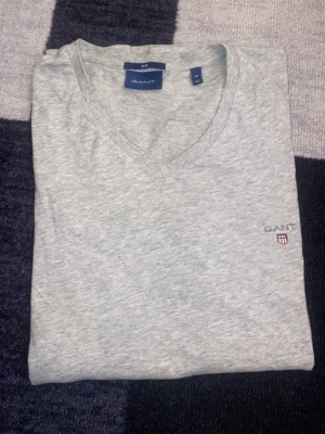 Grå v-ringad t-shirt från Gant - Snygg grå t-shirt från Gant med v-ringning och diskret logga broderad på bröstet. Tillverkad i mjuk bomull som känns skön mot huden. Perfekt basplagg med normal passform och korta ärmar, enkel att matcha till allt.