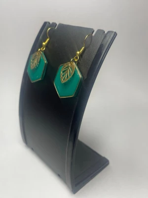 Gröna hexagon örhängen med blad - Snygga örhängen i hexagonformad grön akryl med guldfärgade metalldetaljer och ett litet bladmotiv på varje örhänge. Krokarna är också guldfärgade och ger en cool kontrast till det gröna. Perfekt accessoar för dig som gillar unika former och färger.