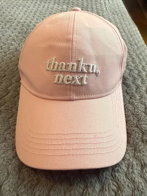 Rosa keps med brodyr 'thank u, next' - Säljer en ljusrosa keps med vit brodyrtext 'thank u, next' framtill. Klassisk böjd skärm och justerbart spänne bak. Tillverkad i bomullstyg, perfekt för en avslappnad och trendig look.