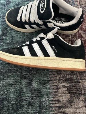 Adidas Campus svarta sneakers - Klassiska Adidas Campus sneakers i svart mocka med vita detaljer och de ikoniska tre ränderna på sidan. Skorna har vit sula med gummiyta undertill och Adidas-logga på plös och häl. Snygg retrostil som passar till många outfits.