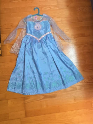 Blå Elsa-klänning från Disney - Snygg blå Elsa-klänning från Disney med glittriga detaljer, transparenta ärmar och snöflingemönster. Perfekt för maskerad eller cosplay. Klänningen har paljettband och skimrande tyg som ger en magisk känsla.