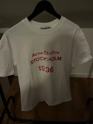 Vit Acne Studios t-shirt med tryck - Säljer en vit t-shirt från Acne Studios med rött tryck framtill där det står 'Acne Studios STOCKHOLM 1996'. Klassisk rund hals och korta ärmar. Perfekt basplagg med snygg streetkänsla och enkel design.