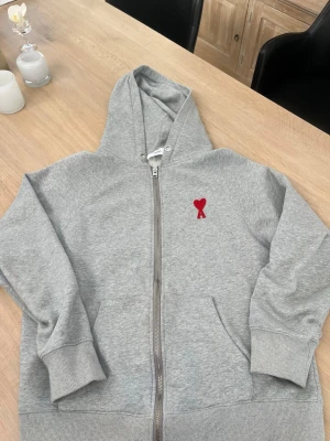 Grå zip hoodie från Ami Paris - Snygg grå hoodie med dragkedja från Ami Paris. Klassisk design med huva, ribbade muddar och fickor. På bröstet sitter Ami:s ikoniska röda hjärta med bokstaven A broderat. Tillverkad i mjuk bomullsblandning för maximal komfort.