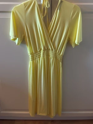 Gul omlottklänning med knyt från Sisters Point - Säljer en ljusgul omlottklänning med korta ärmar och knyt i nacken. Klänningen har resår i midjan för en bekväm passform och är tillverkad i ett mjukt, stretchigt syntetmaterial. Storlek S. Aldrig använd. Från Sisters Point. 