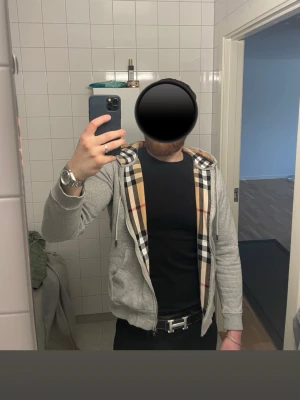 Grå hoodie med Burberry-mönster - Säljer en grå zip-hoodie från Burberry med det klassiska rutmönstret på insidan av huvan och längs dragkedjan. Hoodien har dragsko i huvan, fickor framtill och är tillverkad i mjukt bomullsmaterial. Perfekt för en stilren och avslappnad look.