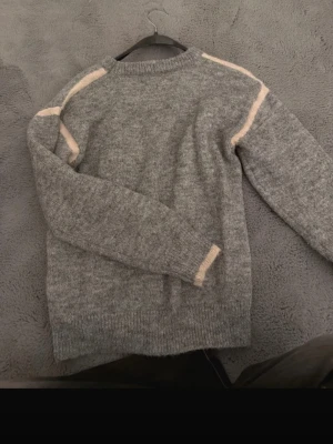 Grå stickad tröja med beige detaljer - Mysig grå stickad tröja med ljusrosa ränder längs axlar och ärmslut. Tröjan har rund halsringning, långa ärmar. Perfekt för kalla dagar och enkel att matcha med olika outfits.