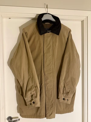 Beige Park Avenue rock från Gant med svart krage - Klassisk beige rock från Gant med svart krage och rutigt innerfoder. Rocken har dold dragkedja och knäppning framtill, samt justerbara ärmslut med knappar. Perfekt för dig som gillar tidlös och stilren design. Väldigt liten fläck som säkert går bort med en tvätt, annars typ nyskick! 