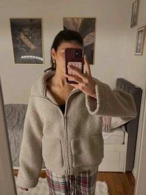 Beige teddyjacka  - Mysig beige teddyjacka med hög krage och dragkedja framtill. Jackan har två stora fickor och är gjord i fluffigt teddy-material som håller dig varm. Perfekt oversized passform för en avslappnad look.
