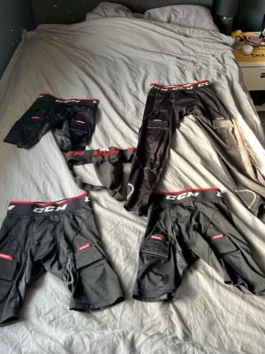 Svarta hockeyshorts och skydd från CCM - Fyra par svarta hockeyshorts och ett suspensoar från CCM. Shortsen har röda och vita detaljer, elastisk midja med CCM-logga och fickor på sidorna. Materialet är slitstarkt och anpassat för sport. Perfekt för hockeyträning eller match.