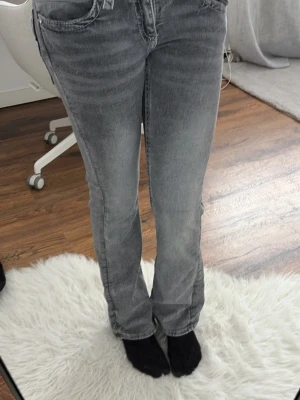 Grå raka jeans med detaljer - Säljer ett par grå jeans med raka ben och snygga slitningar. Jeansen har dekorativa bakfickor med flätade kanter och silverfärgade knappar. Perfekta för dig som gillar en avslappnad men ändå trendig look.