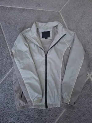 Ljusgrå vindjacka från Zara med vita detaljer - Stilren ljusgrå vindjacka med vita kontrastsömmar och svart dragkedja framtill. Jackan har hög krage, elastiska muddar vid ärmar och nederkant samt meshfoder på insidan för extra komfort. Perfekt för blåsiga dagar och enkel att matcha med olika outfits.
