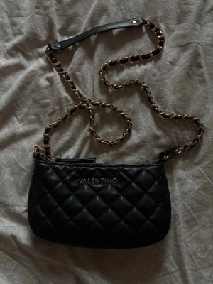 Svart quiltad axelväska Valentino - Snygg svart axelväska från Valentino med quiltat mönster och guldfärgad kedjerem med läderdetalj. Väskan har en stilren, rektangulär form och dragkedja upptill. Perfekt för dig som gillar trendiga accessoarer med lyxig känsla.
