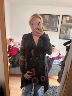 Desigual kofta - Unik svart kofta med färgglada mandala-liknande mönster i rött, grönt, gult och orange. Koftan har lång ärm, öppen framtill och asymmetrisk nederkant. Perfekt för dig som gillar boho-stil och vill sticka ut med coola detaljer.