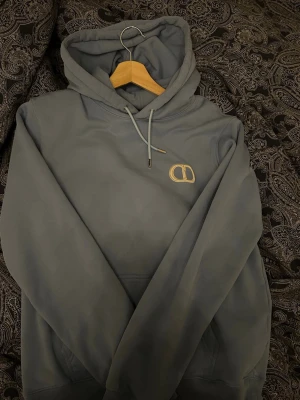 Grå hoodie med CD-logga - Säljer en stilren grå hoodie med huva och dragsnören. På bröstet finns en broderad CD-logga i beige. Hoodien har ribbade muddar vid ärmslut och nederkant samt en klassisk känguruficka framtill. Perfekt för dig som gillar enkel och snygg design.