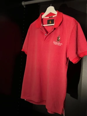 Rosa pikétröja från Polo Club, Royal County of Berskshire version - Den ända Royal County of Berkshire tröjan på plick , Och svår att hitta på nätet med just den färgen och dessa broderingar. Säljer en snygg rosa pikétröja från Polo Club (Royal County of Berkshire) med broderad logga på bröstet och tryck på ärmen. Klassisk krage och korta ärmar, perfekt till sommaren. Tillverkad i mjuk bomull för skön känsla. 