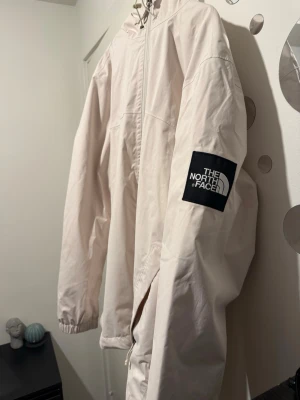 Ljusbeige vindjacka från The North Face - Snygg ljusbeige vindjacka från The North Face med huva och dragkedja framtill. Jackan har elastiska muddar vid ärmsluten och en tydlig logga på ärmen samt ryggen. Perfekt för blåsiga dagar och enkel att matcha med olika outfits.
