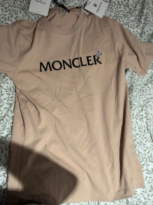 Beige Moncler t-shirt med logga - Snygg beige t-shirt från Moncler med stor svart logga framtill och klassisk rund hals. T-shirten har korta ärmar och är tillverkad i mjuk bomull, perfekt för en clean och stilren look.