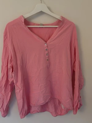 Rosa blus från Esprit med knappar - Snygg rosa blus från Esprit med v-ringning och dekorativa knappar framtill. Blusen har lång ärm och är tillverkad i ett tunt, mjukt tyg som ger en avslappnad look. Perfekt att styla med jeans eller kjol för en trendig outfit.