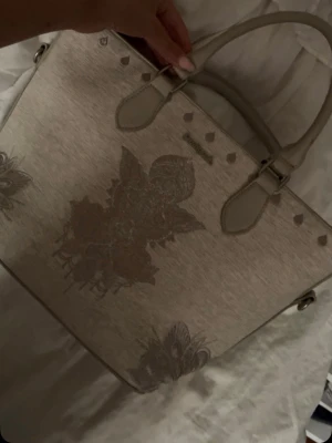 Beige handväska från Desigual - Snygg beige handväska från Desigual med dekorativa blommönster och små hjärtan. Väskan har två rejäla handtag och detaljer i samma färg som väskan. Materialet ser ut att vara konstläder med en matt finish. Perfekt för dig som gillar unika detaljer och stilren design.
