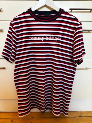 Randig Tommy Hilfiger t-shirt storlek S herr - Säljer en randig t-shirt från Tommy Hilfiger med broderad logga på bröstet. T-shirten har marinblå, röda och vita ränder, rund halsringning och korta ärmar. Perfekt skick! Bomull, storlek S herr