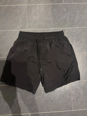 Svarta shorts med snörning - Säljer ett par svarta shorts med elastisk midja och snörning framtill. De har en enkel och clean design, perfekta för sommardagar. Materialet känns lätt och bekvämt, vilket gör dem smidiga att röra sig i.